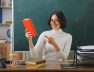 smiling-young-female-teacher-wearing-glasses-holding-points-book-sitting-desk-with-school-tools-classroom