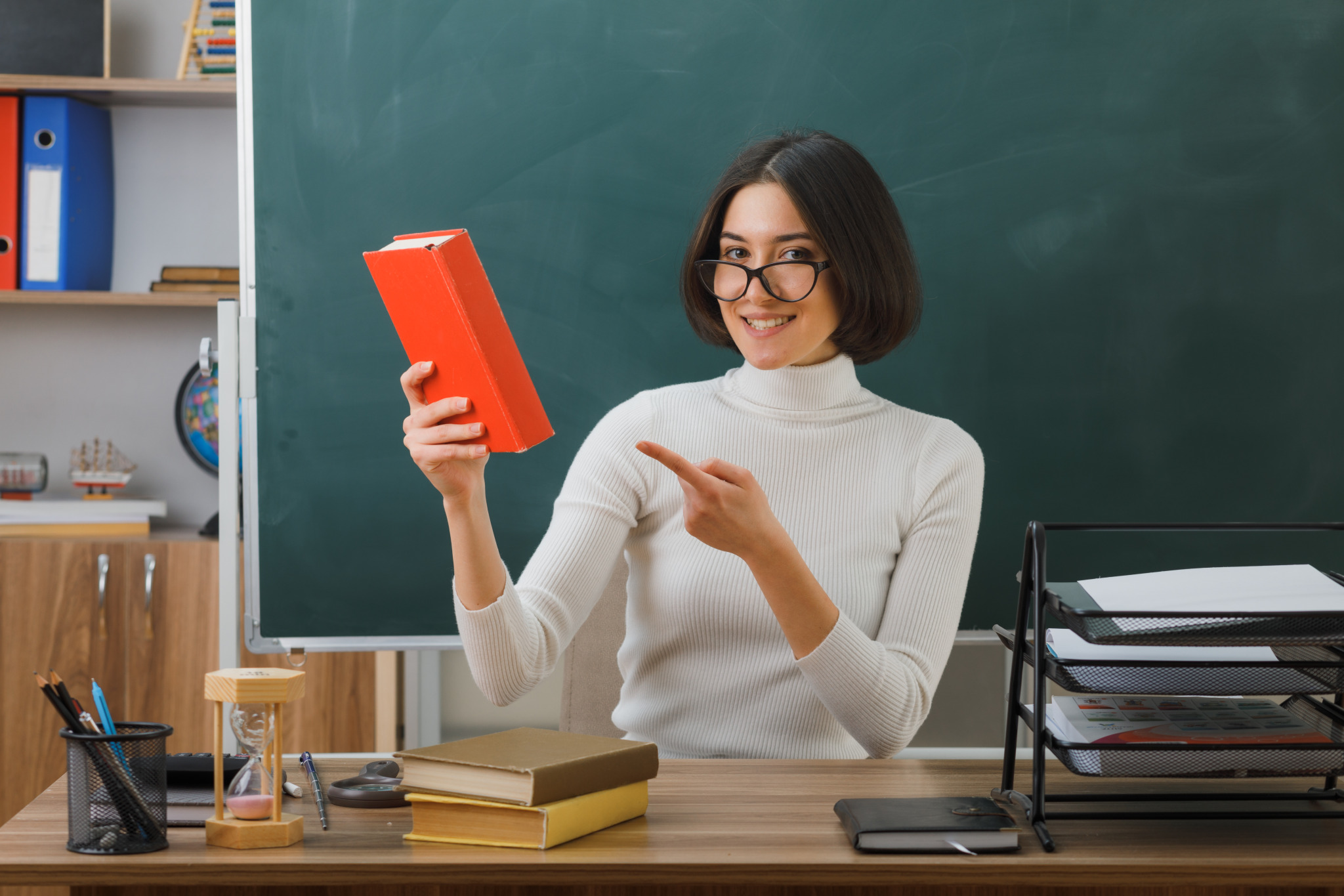 smiling-young-female-teacher-wearing-glasses-holding-points-book-sitting-desk-with-school-tools-classroom